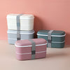 Quatre Lunchbox en plastique à deux compartiments posé sur un fond beige et rose. Il y a une lunch box blanche, une grise, une verte et une rose.