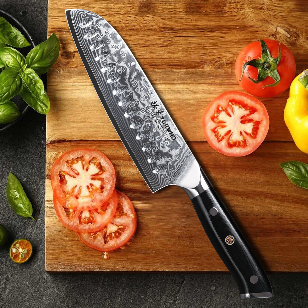 Couteau santoku de style japonais en acier de Damas posé sur une planche en bois avec des tomates coupées à gauche et à droite sur fond gris