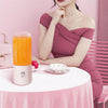 Blender portable pratique pour fruits et légumes posé sur une table rose avec un bol et un sac à main rose, accompagné d'une femme portant une robe rose sur fond rose