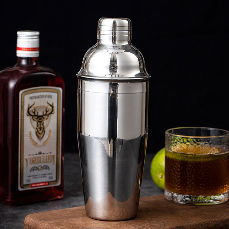 Shaker à cocktail en acier inoxydable sur une planche en bois avec une bouteille d'alcool à gauche et un verre rempli d'alcool à droite à côté d'un citron vert sur fond noir