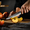 Couteau de cuisine japonais en acier de Damas avec manche noir en train de couper une pêche sur une planche en bois dans les mains d'un homme sur fond gris