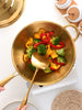 Sauteuse dorée avec poignée en acier inoxydable posée sur une table blanche avec une spatule et des légumes à l'intérieur