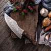 Couteau de boucher viking en acier inoxydable avec manche en bois posé sur une table avec des brindilles en haut, du tissu et des légumes sur la droite