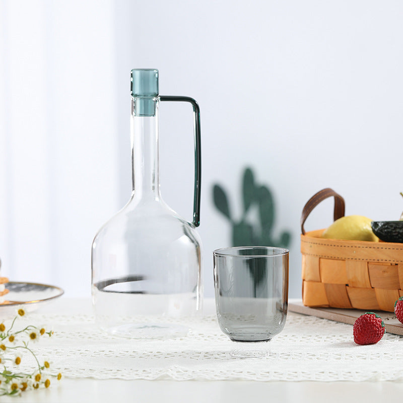 Carafe d'eau en verre coloré nordique posée sur une table grise avec un verre et un panier en osier à droite sur fond blanc